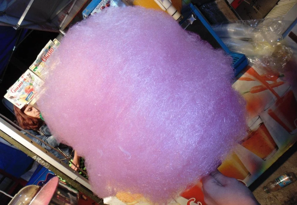 A hand holding a stick of pink/purple candyfloss against an urban-seeming background