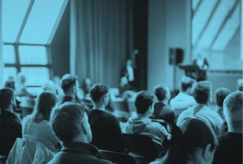 A group of people seated in a conference or lecture room, facing a speaker at the front near a podium.