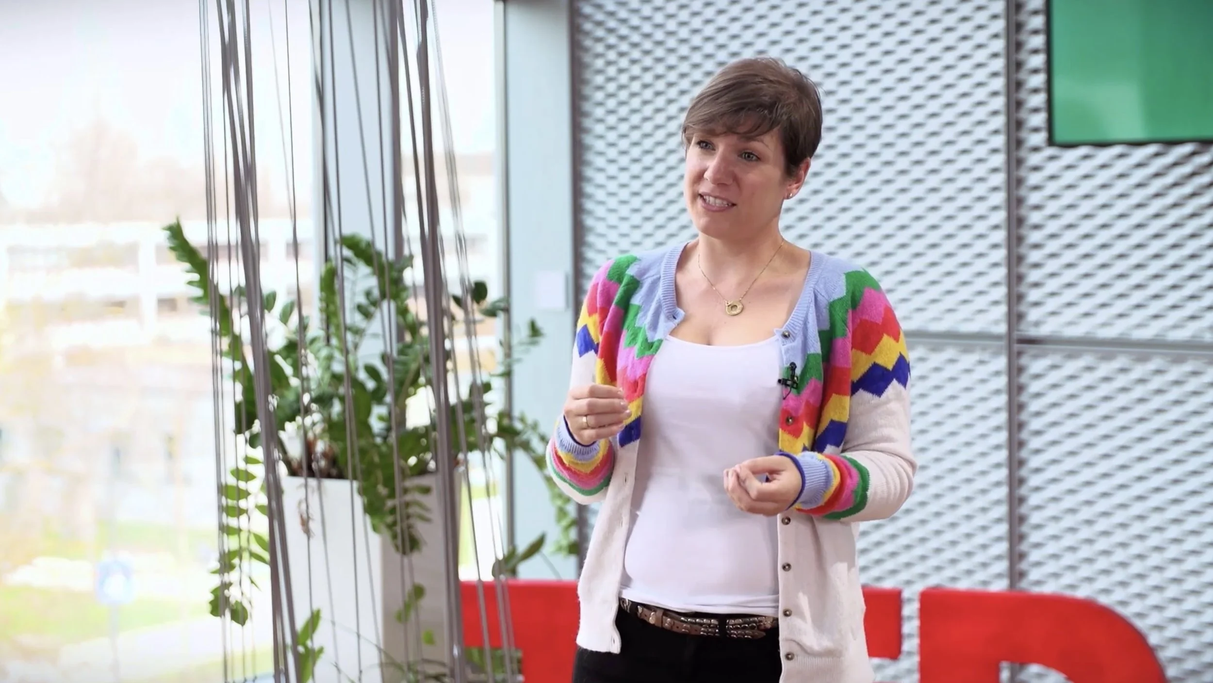 Alexandra Wander speaking during the TEDxTUMStudio recording on 9 April 2021 at TU Munich, wearing a colourful zig-zag cardigan, hands mid-gesture, red TEDx letters visible in the background. She's discussing why it takes a higher level of autonomy.