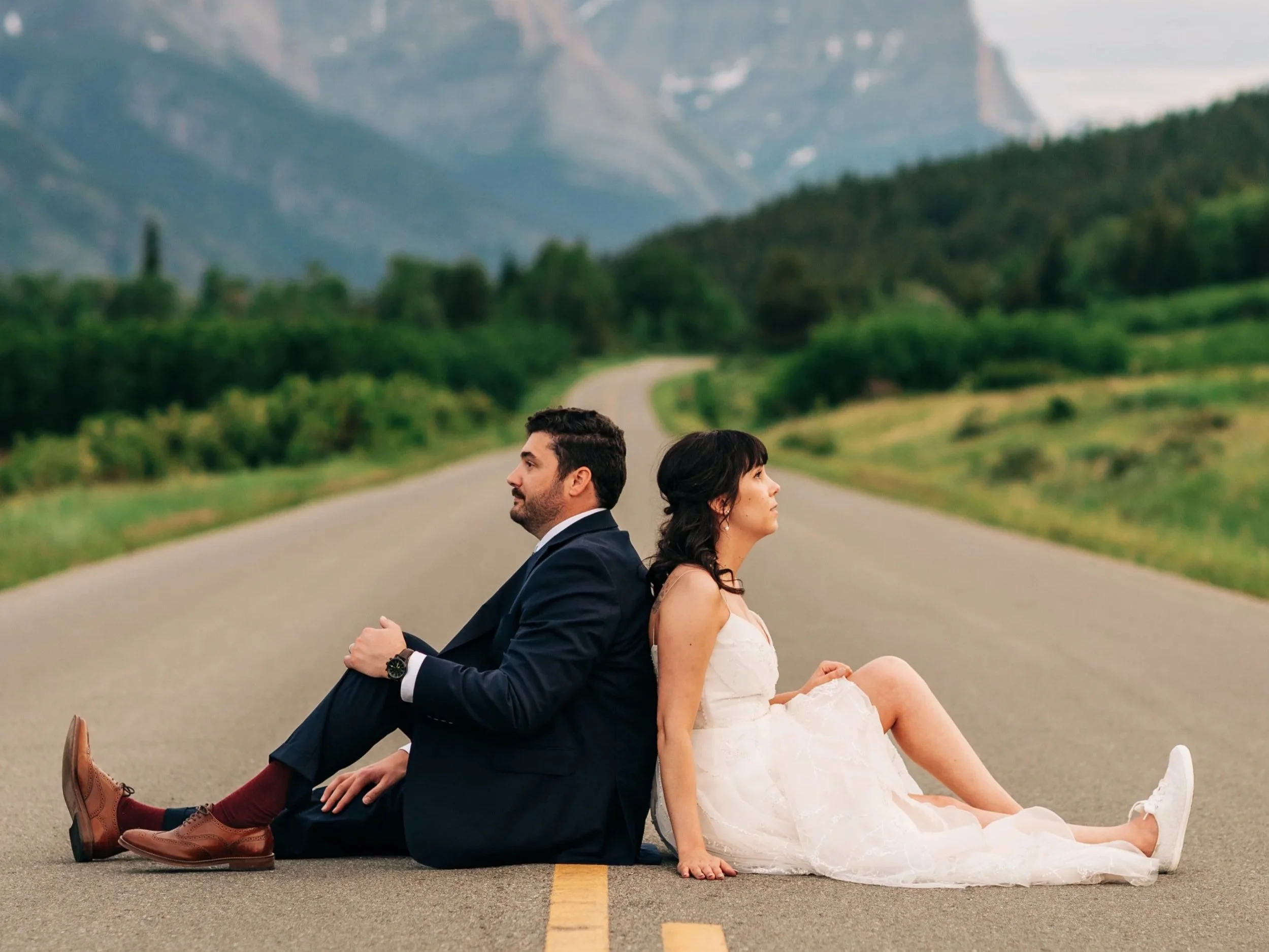 A couple in wedding attire standing in a field of purple flowers with mountains in the background at sunset.