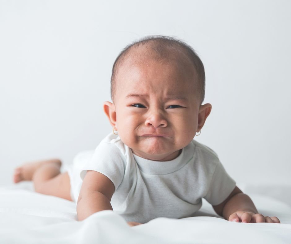 A baby wearing a white vest lying on stomach with head raised looking at camera - crying