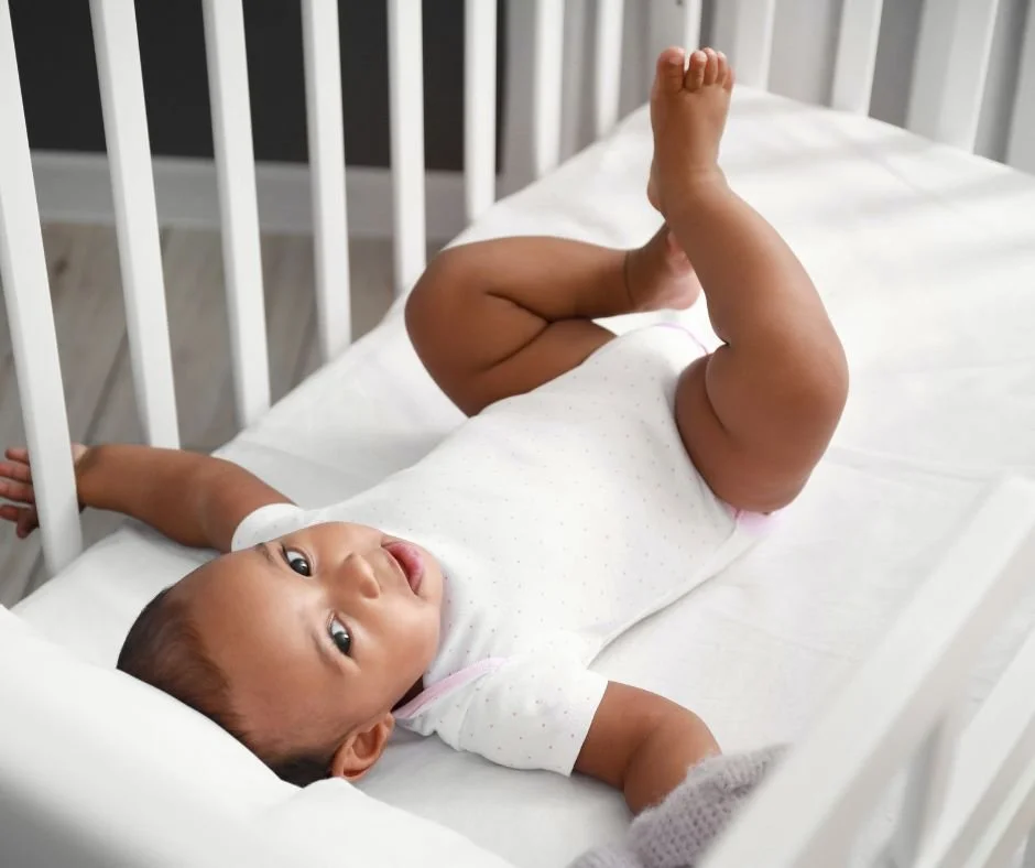 Baby lying in the cot with one leg kicked in the air, wide awake and alert instead of settling to sleep