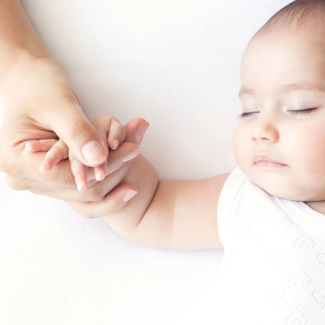 Baby in white vest asleep with adult hand holding baby's hand