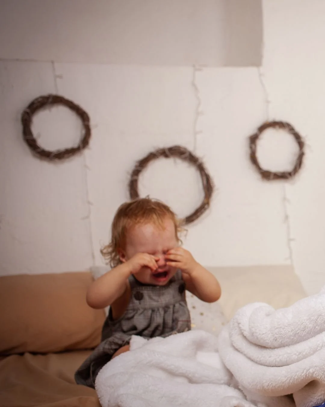 Toddler sitting on bed with whie blanket, rubbing here eyes and visibly upset
