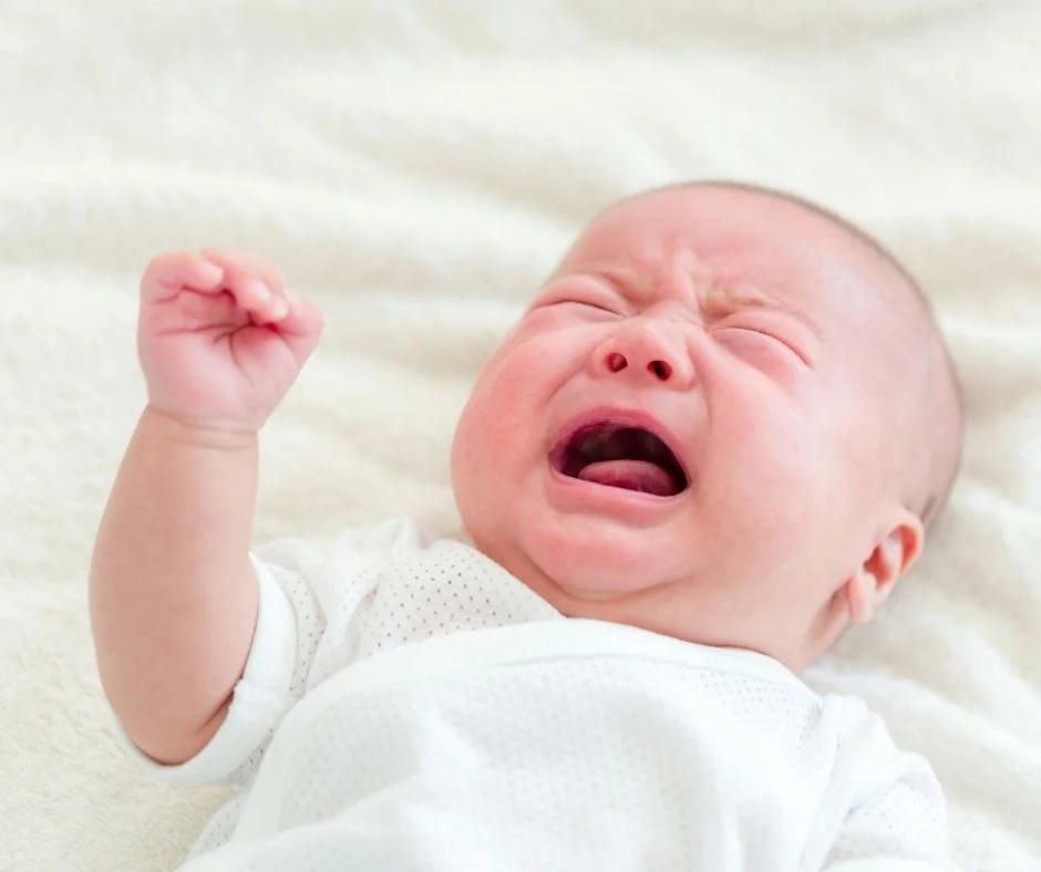 Baby wearing white lying on back crying with one arm raised