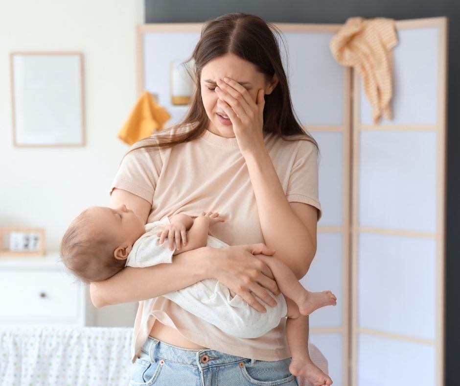 Exhausted mum holding baby with her hand up against her forehead
