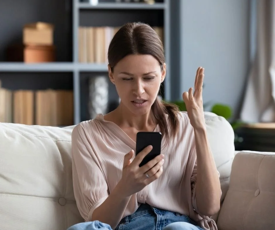 Woman sitting on sofa holding a phone with the other hand held up looking  confused and exasperated