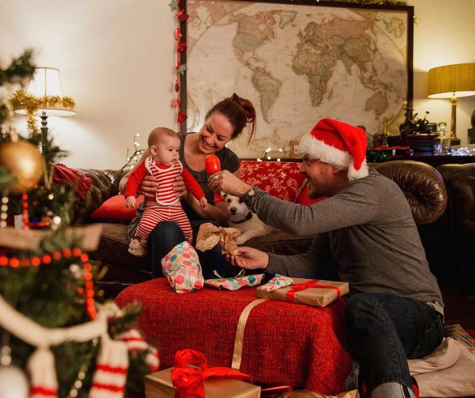 Romm decorated for Christmas with  a mum holding baby on her lap and dad wearing Santa hat showing baby a large red rattle.