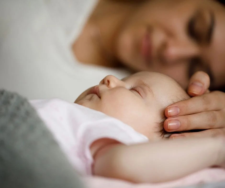 Sleeping baby sharing bed with mum who's lying alongside with her hand on baby's head