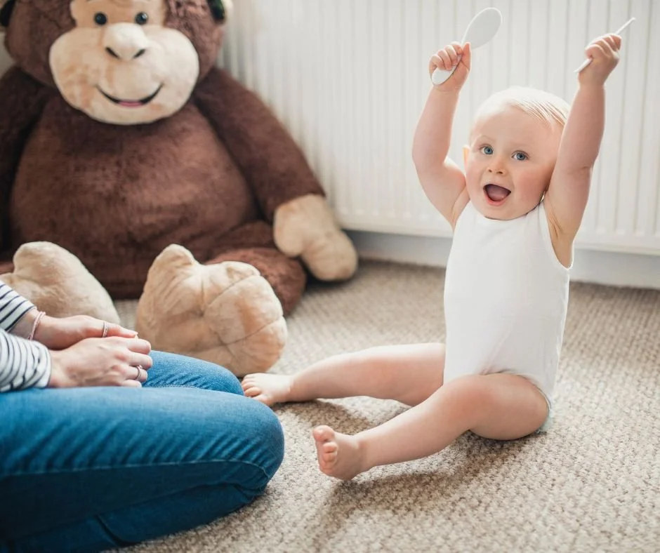 Baby sitting on the floor with arms raised high and a big open-mouthed grin, wired with energy instead of winding down