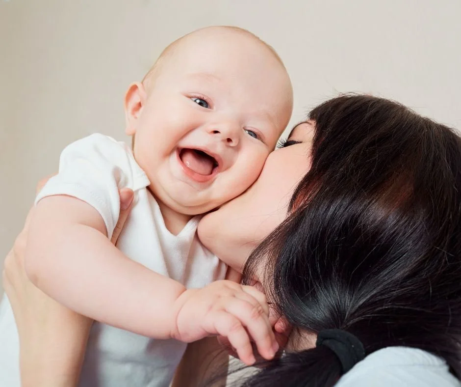 Very smiley baby being help up by mum, and mum, with long dark hair, kissing baby on cheek