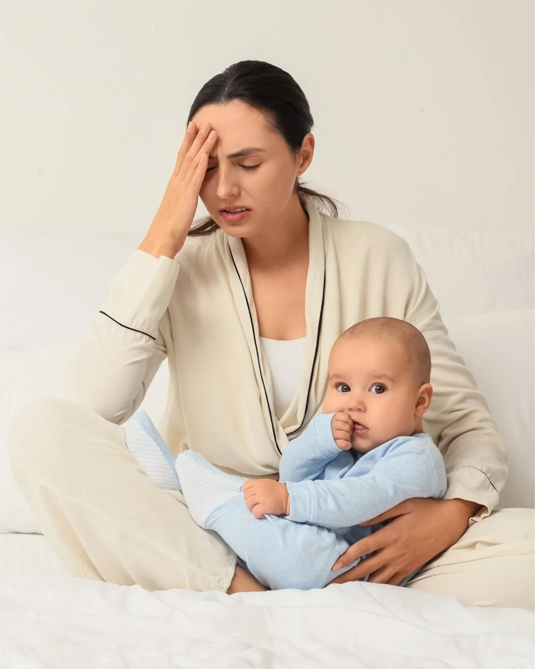 Mum holding her hand to her forehead, eyes closed looking stressed with a baby wearing a blue sleepsuit siting on her lap