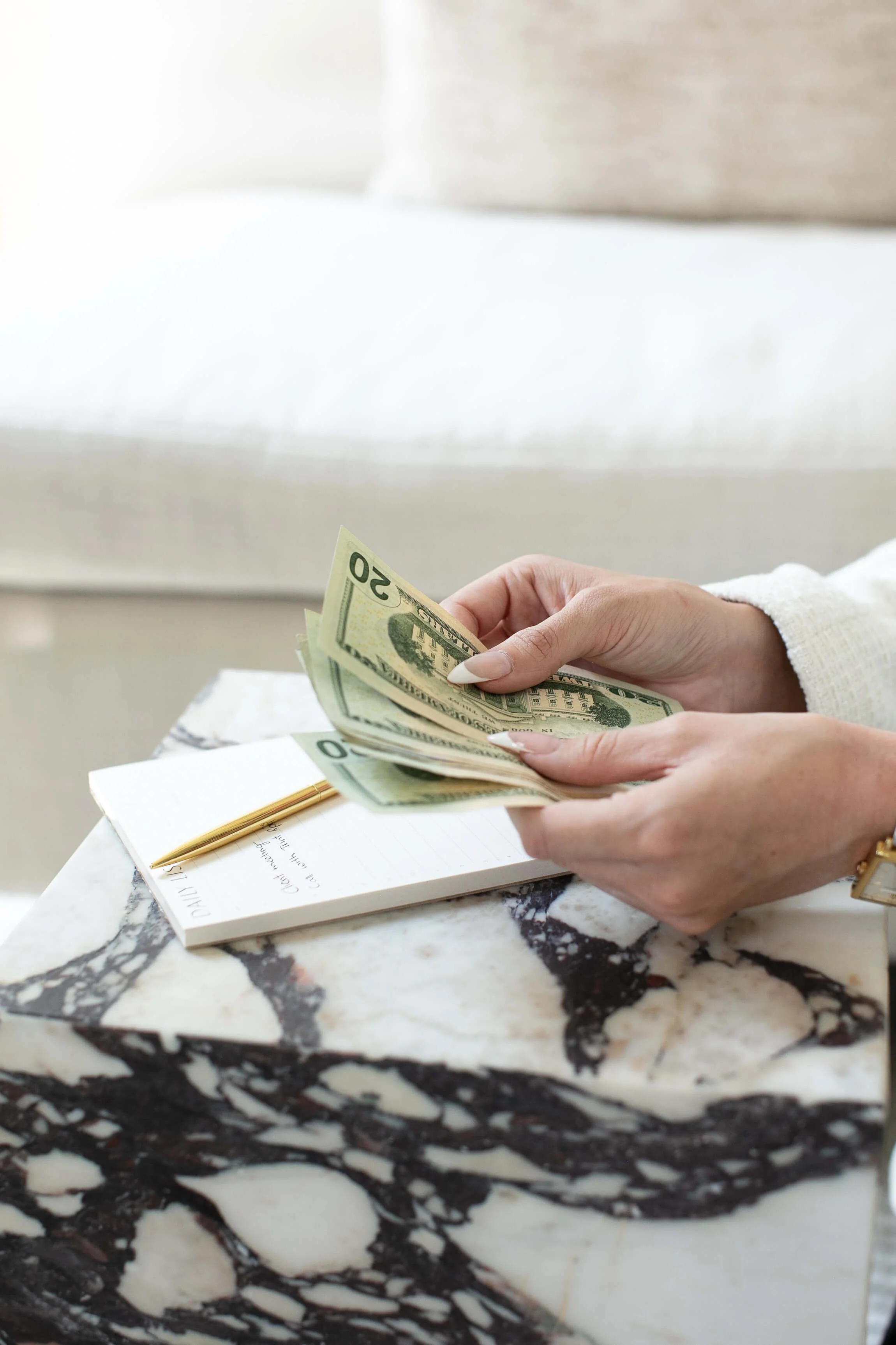 Person holding multiple twenty-dollar bills while seated at a marble table, with a notebook and gold pen nearby.