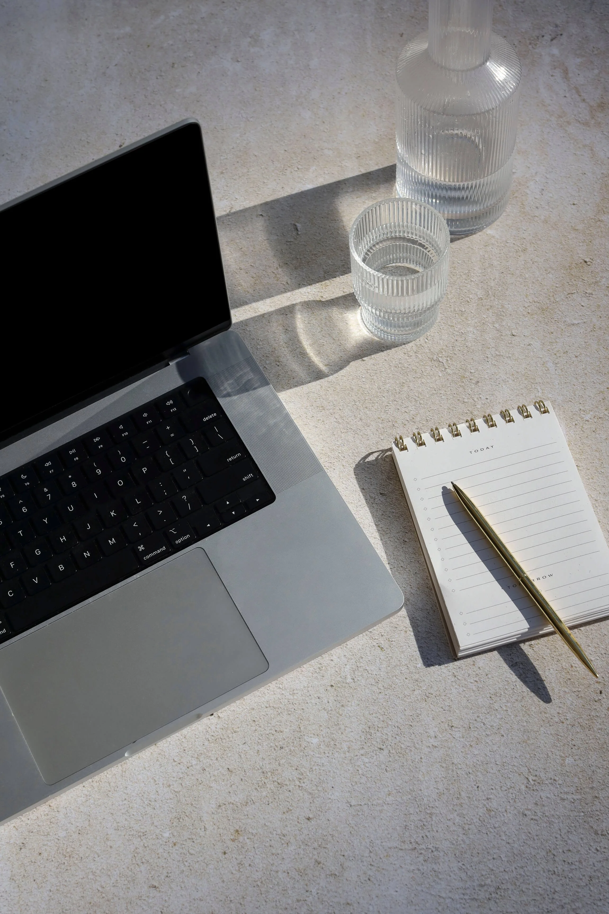 A silver MacBook on a white textured surface, next to a spiral notepad with a gold pen, a glass water bottle, and a glass of water. Bright sunlight casts shadows.