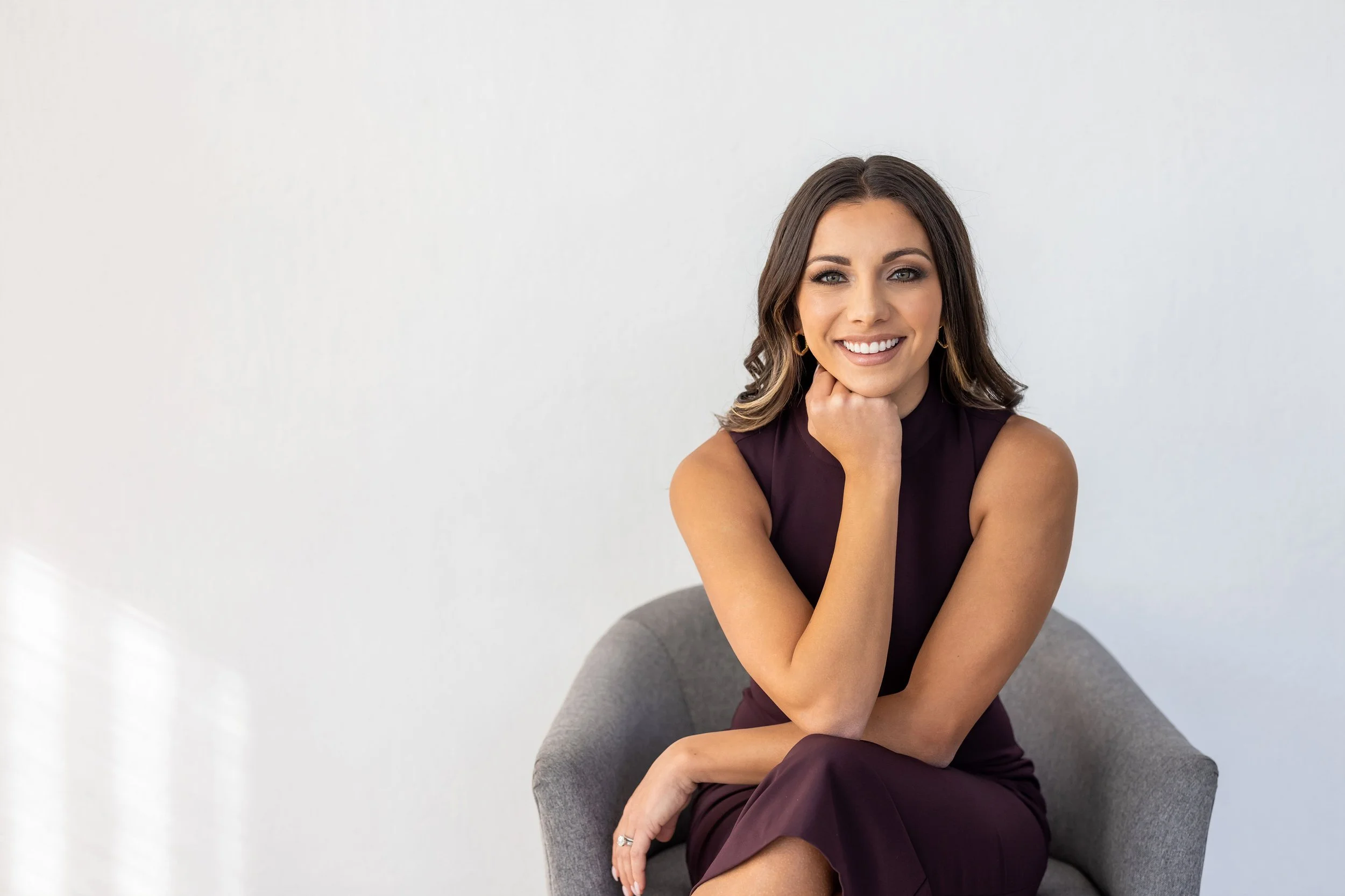 A woman in a sleeveless dark purple dress sitting on a gray armchair, smiling with her chin resting on her hand, against a plain white background.