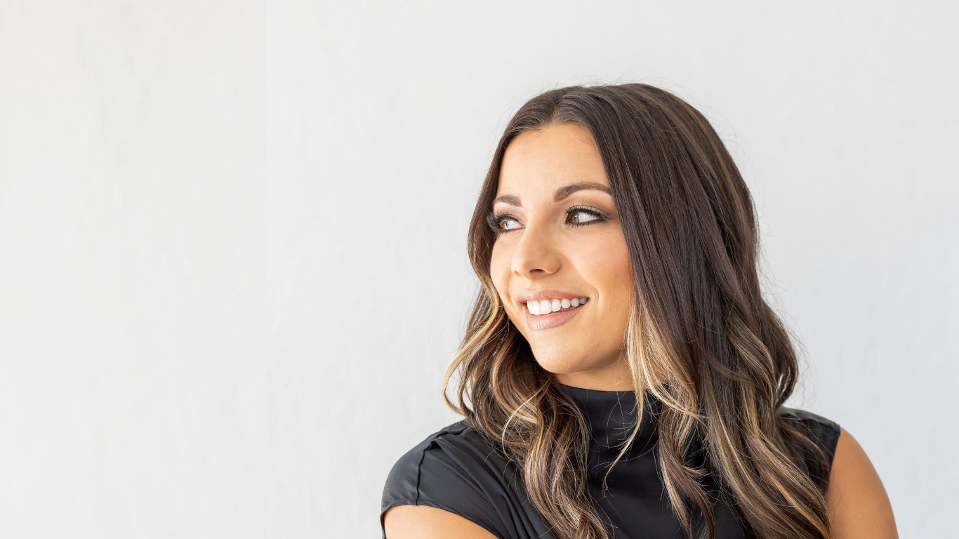 A young woman with wavy brown hair and a black sleeveless top, smiling and looking to her left, standing against a plain light-colored wall.