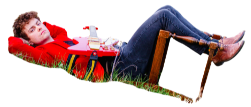 A young boy is lying on grass with his feet elevated on a wooden chair. He is wearing a red sweater and jeans, damien alblas the musician.