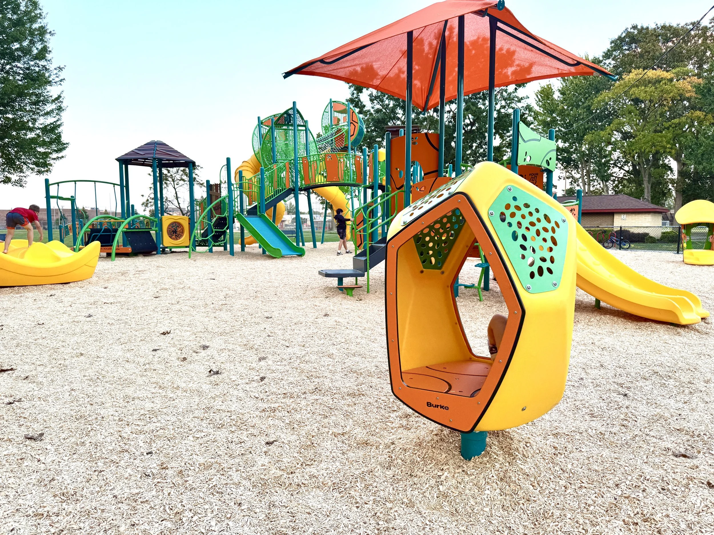 Bri yellow, orange and green playground with a fun spinning play piece in the middle, large curly slides and a big shaded umbrella
