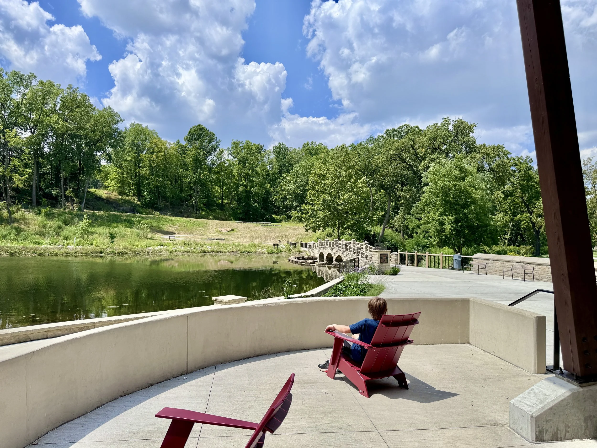 Photo shows a child sitting in a red chair over looking a beautiful lake with a cobblestone bridge