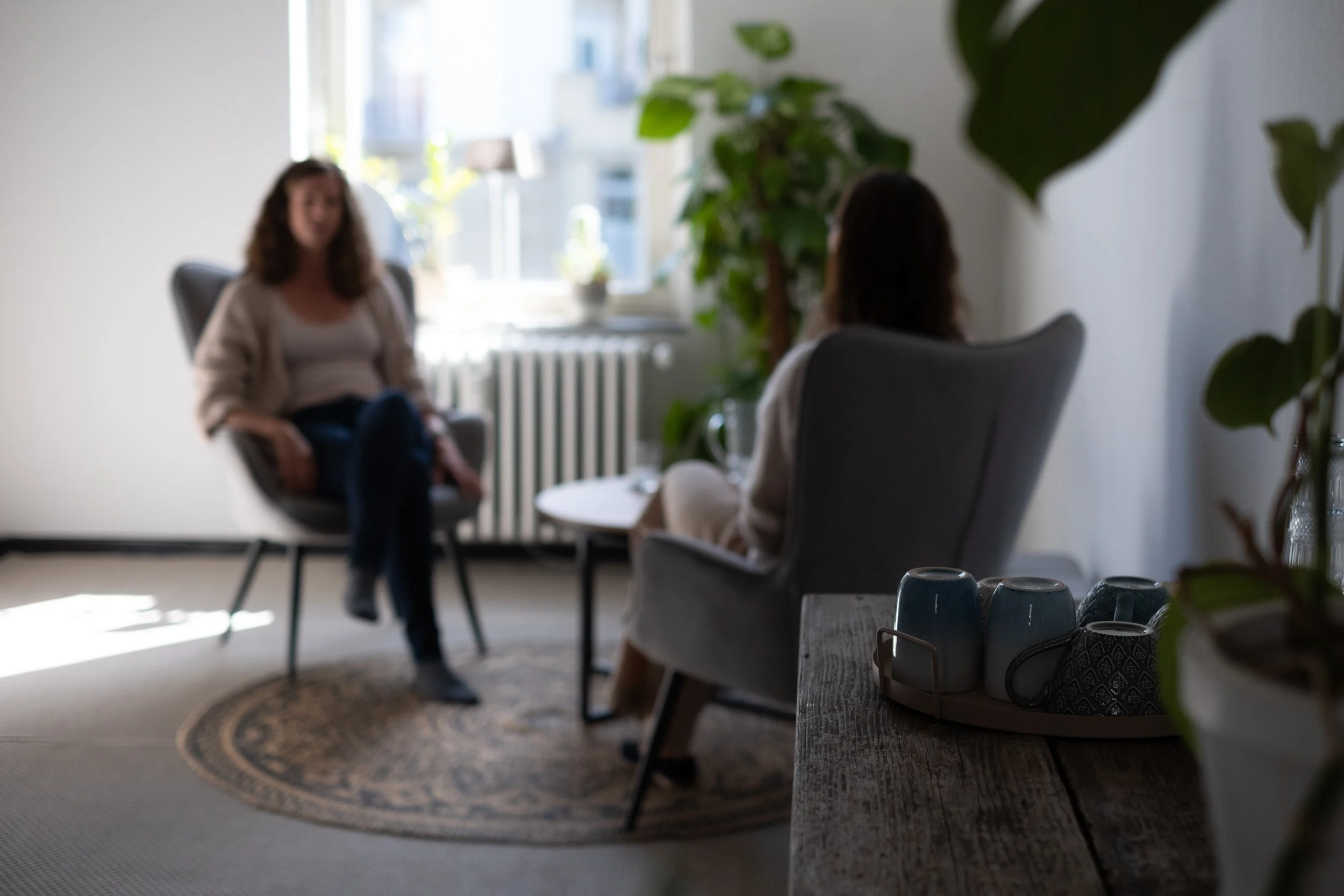 Sarah Graf sitzt mit ihrer Klientin auf zwei großen gemütlichen Sesseln. Im Vordergrund sieht man den rustikalen Landhaustisch mit der Tee- und Kaffeeecke. Auf dem Boden liegen gemütliche Teppiche. Im Hintergrund scheint die Sonne durchs Fenster.