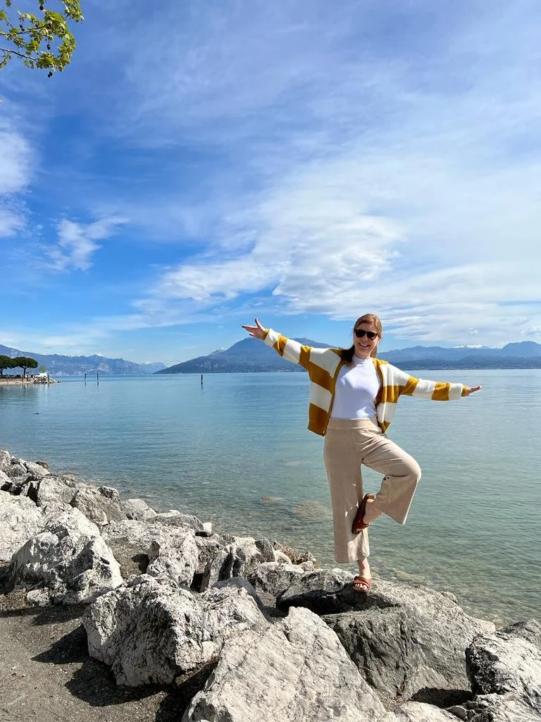 Frau steht am See auf einem Bein und hält die Balance. Ihre Arme sind beide weit ausgestreckt. Sie lacht, die Sonne scheint.