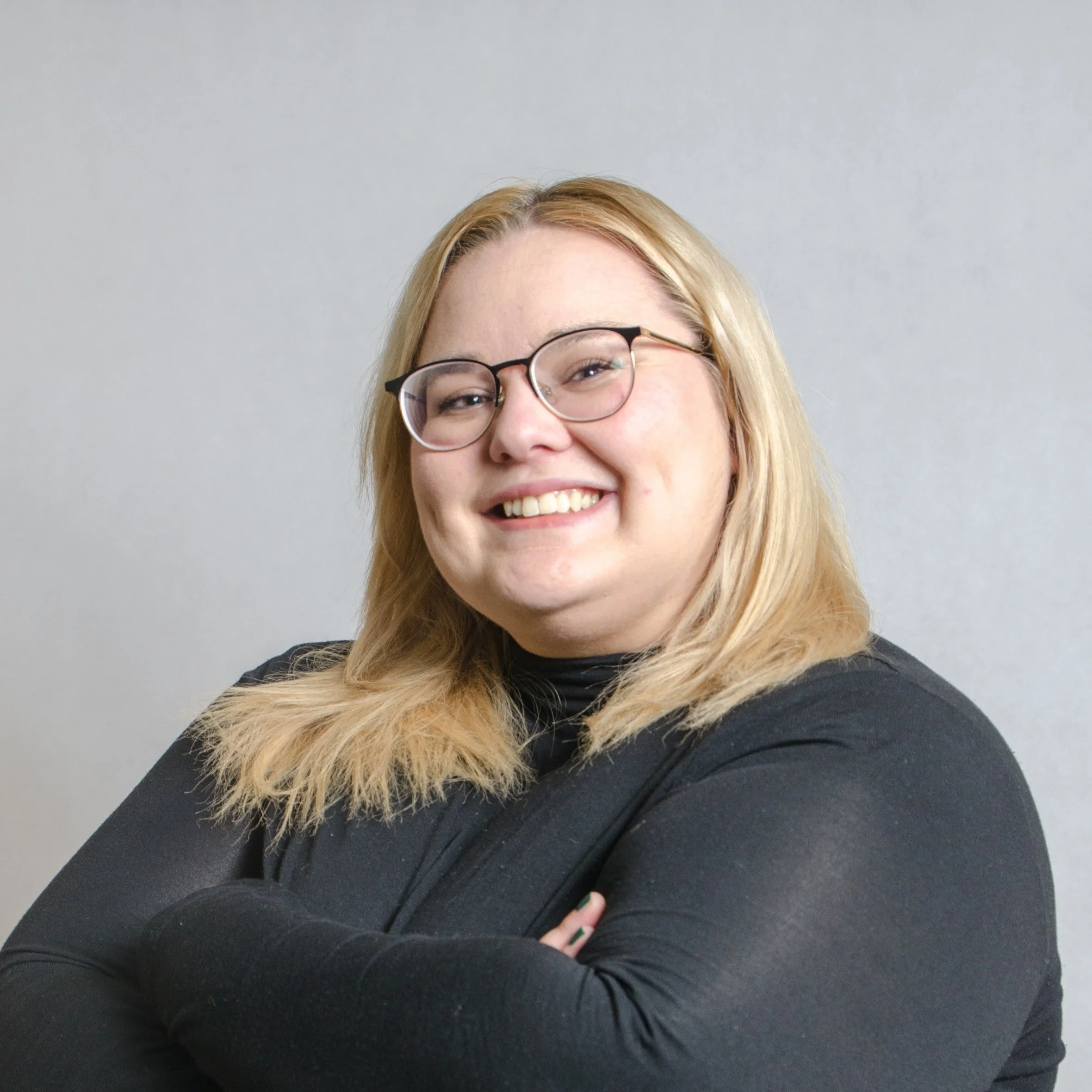 Blonde korpulente Frau lächelt seitlich in die Kamera. Sie trägt einen schwarzen langen Pullover und wirkt sehr zuversichtlich. Der Hintergrund ist schlicht grau.