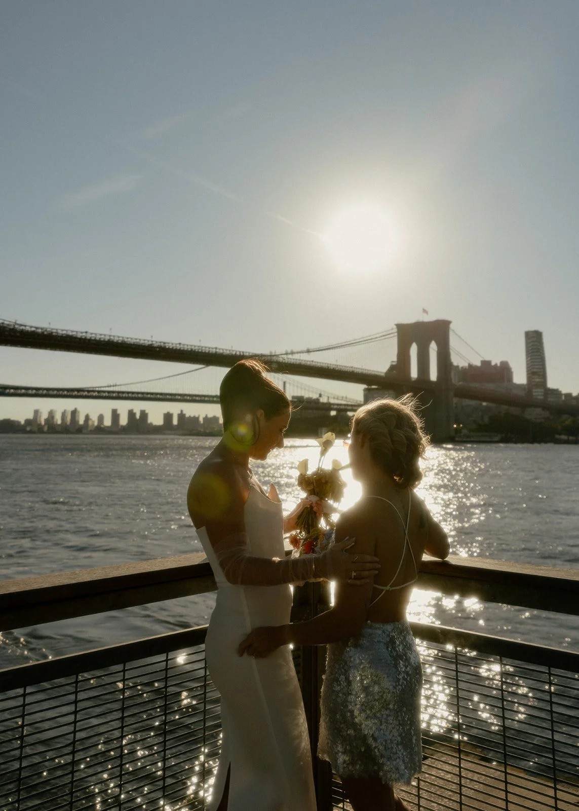 LGBT brides admiring the Brooklyn Bridge from afar as the sun rises and shines over the river.