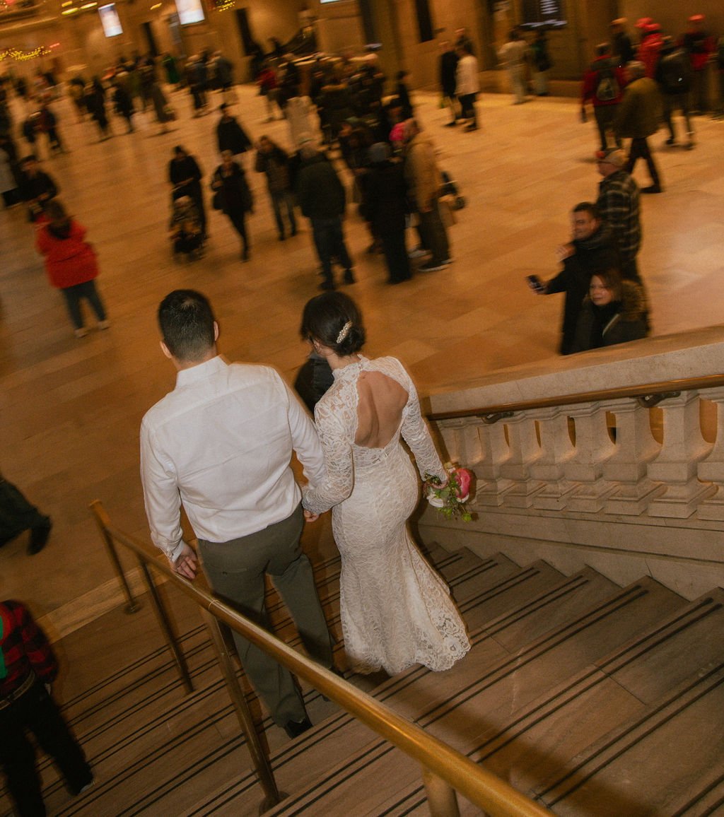 A bride and groom holding hands walking down a staircase amid a crowd at what appears to be a celebration or wedding reception.