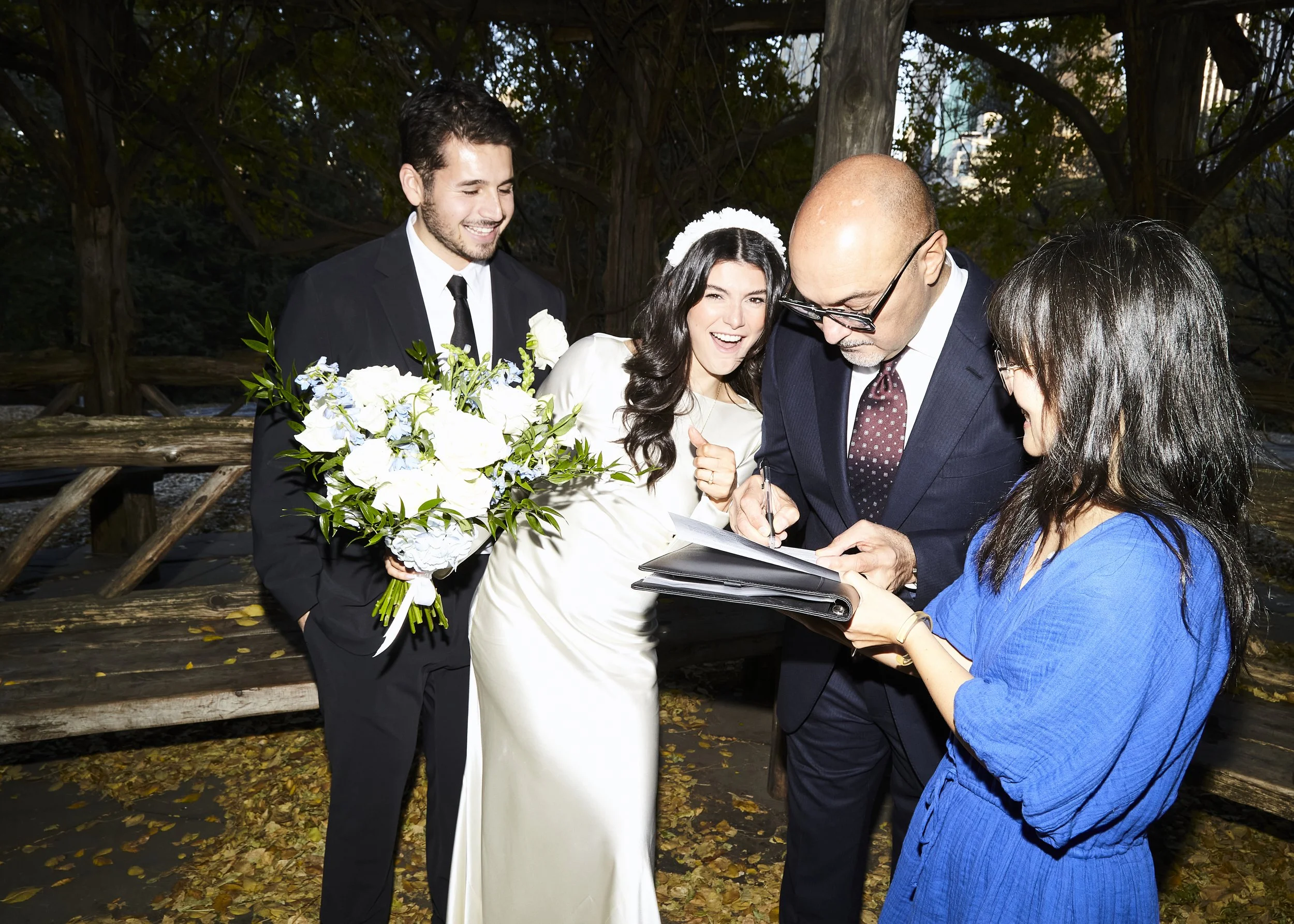 Bride and groom smiling while their witness and officiant sign their marriage license during their Cop Cot wedding ceremony at Central Park, planned by Cakewalk.