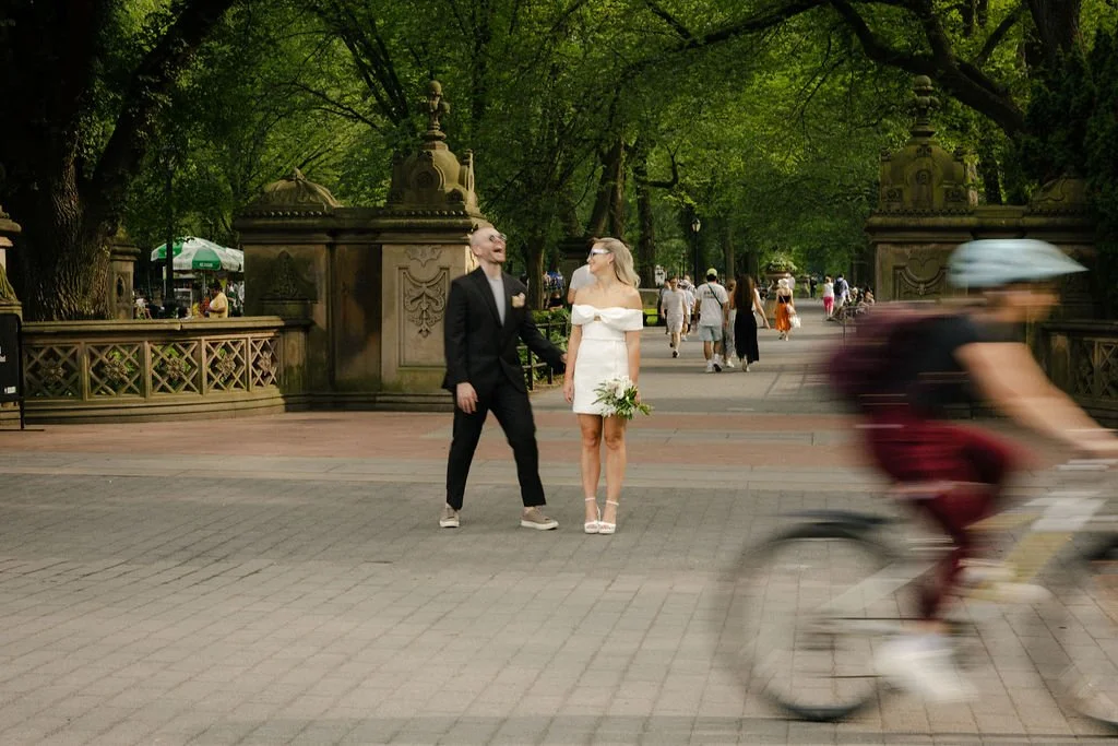 Bride and groom laughing in Central Park with a biker whirring by, on the way to their Cakewalk elopement.