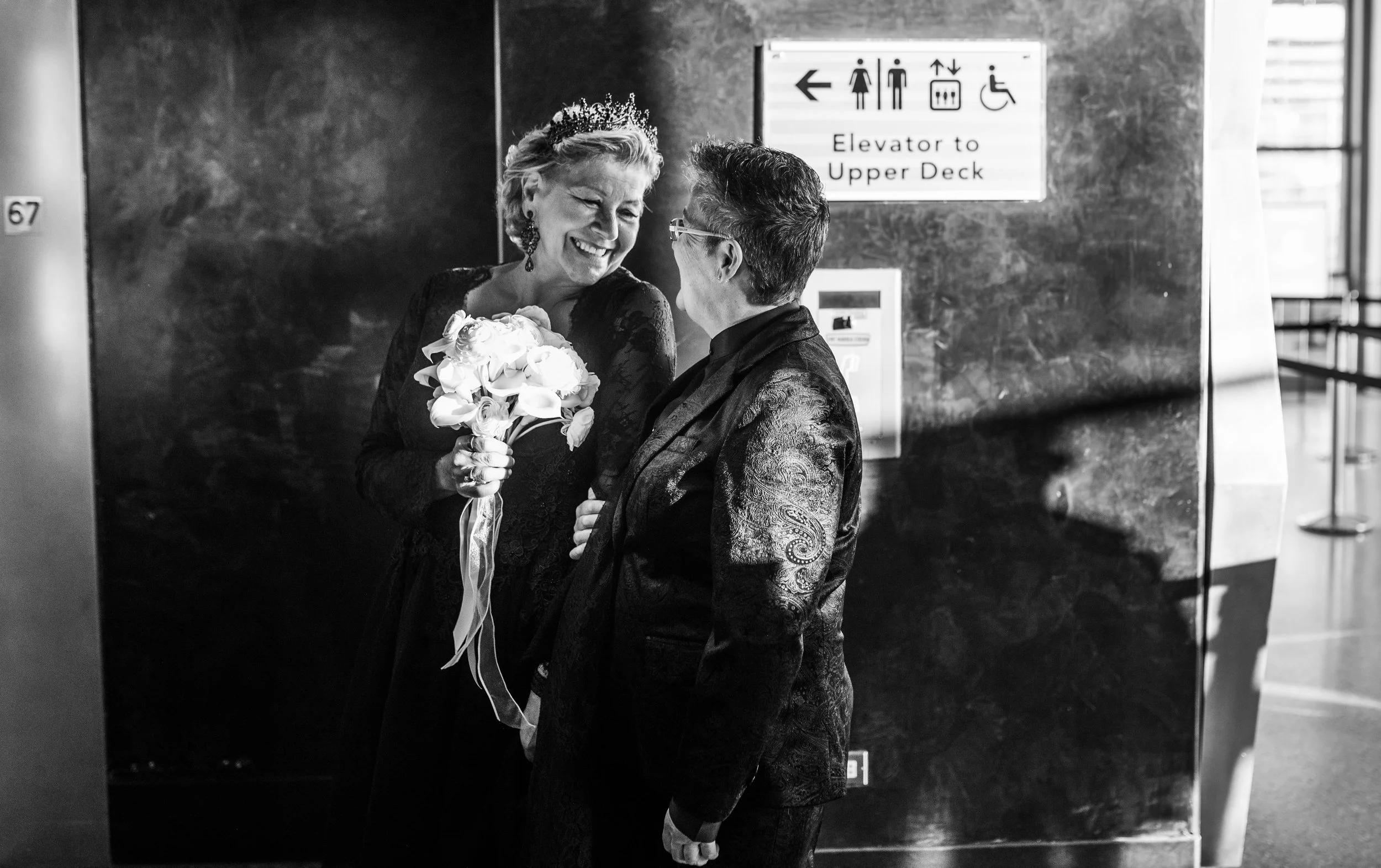 Two women face each other smiling before entering the elevator at the Top of the Rock on the way to their Cakewalk elopement.