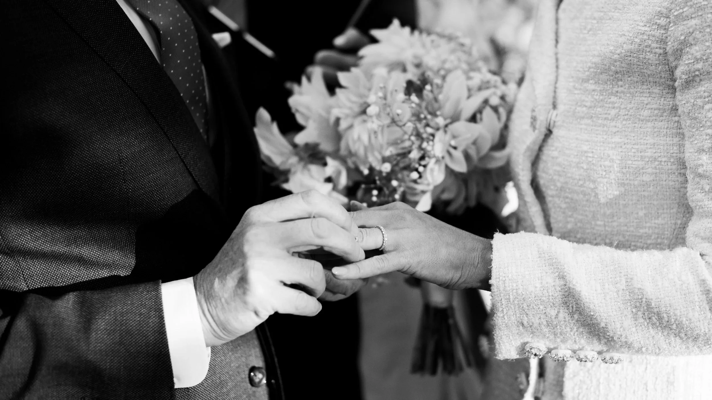 Close-up of a wedding ceremony where a groom is placing a ring on the bride's finger. The bride's hand has a wedding ring, and the groom is wearing a suit and polka dot tie. The bride is holding a bouquet of flowers.
