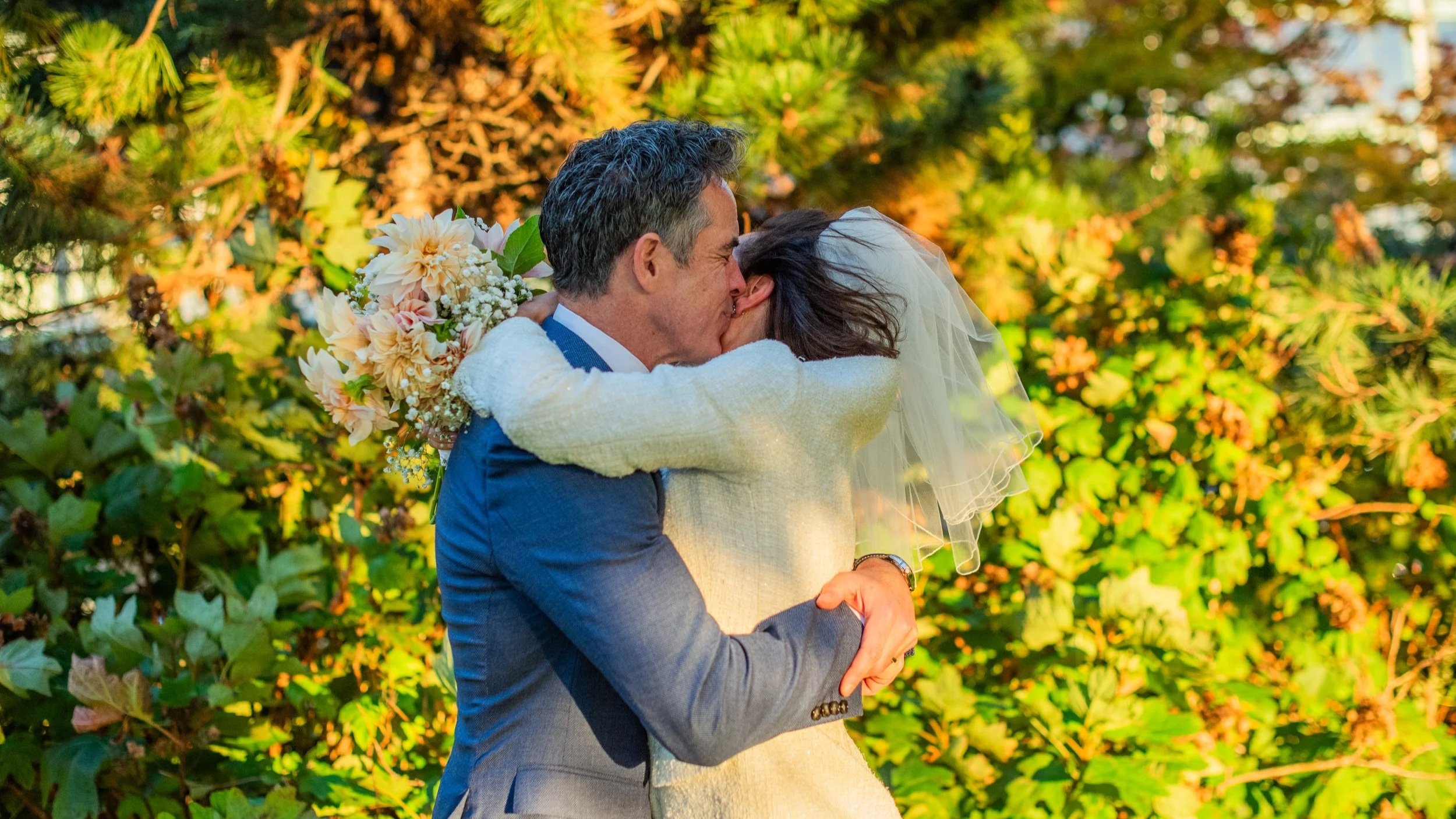 A couple sharing a kiss outdoors, the man in a blue suit and the woman in a white sweater and veil, with green foliage in the background.