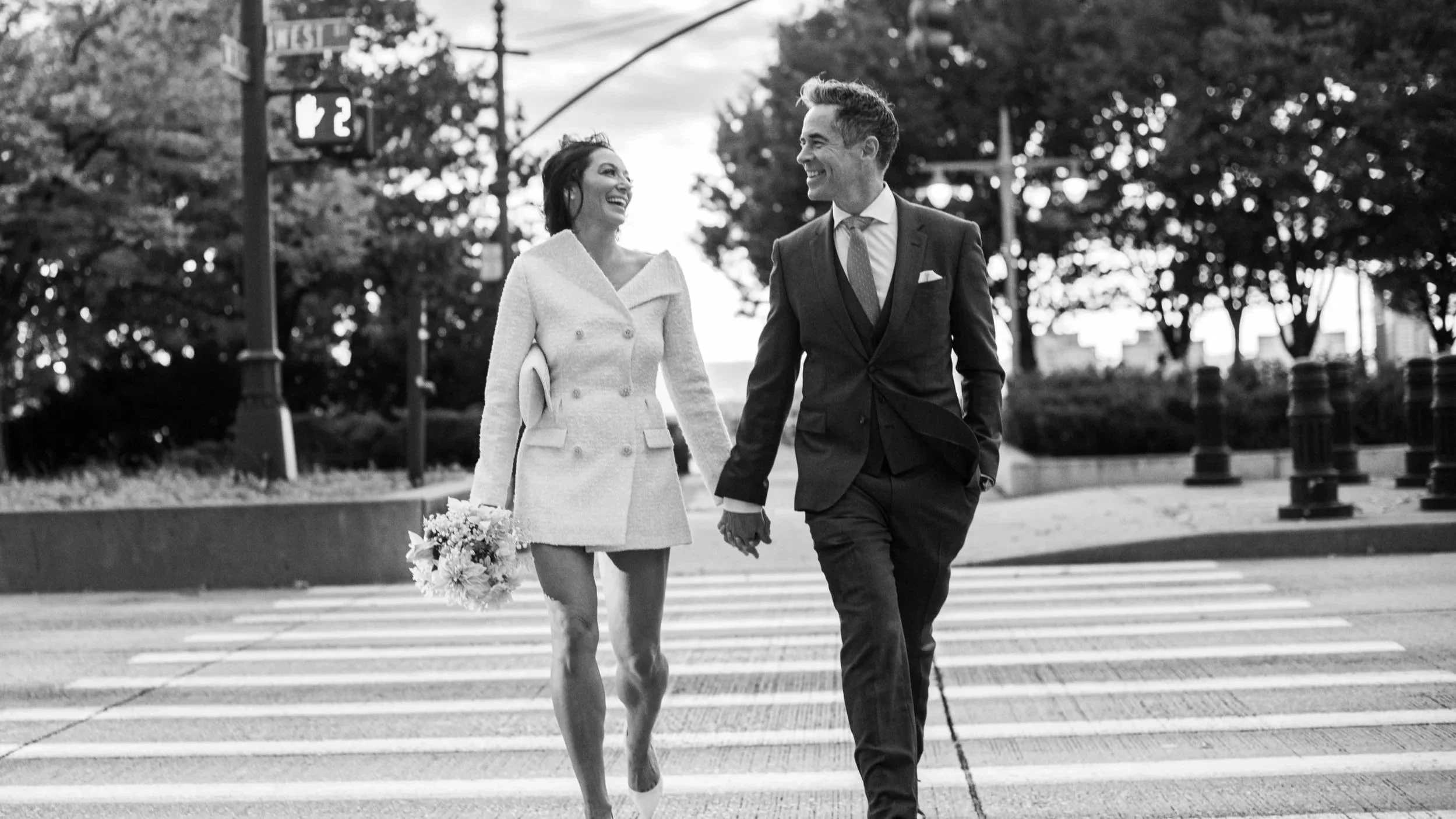 A smiling woman and man holding hands and walking across a crosswalk in a city park, with trees and streetlights in the background.