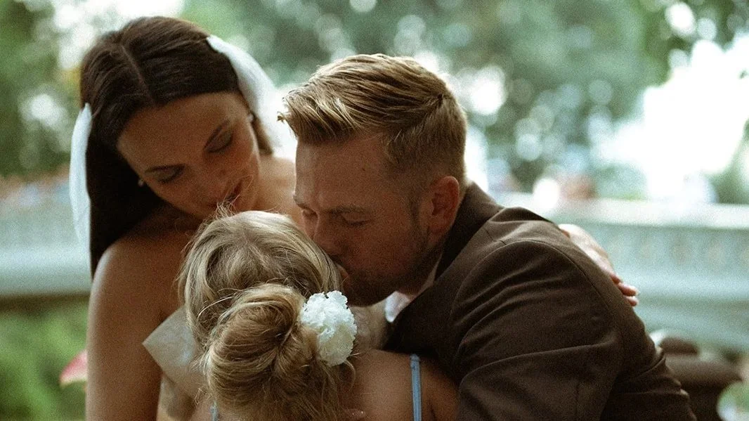A woman, a man, and a young girl are sharing a warm embrace outdoors. The woman has long dark hair with a white ribbon, and the girl has blonde hair styled in two buns with white flowers. The background shows blurred trees and outdoor scenery.
