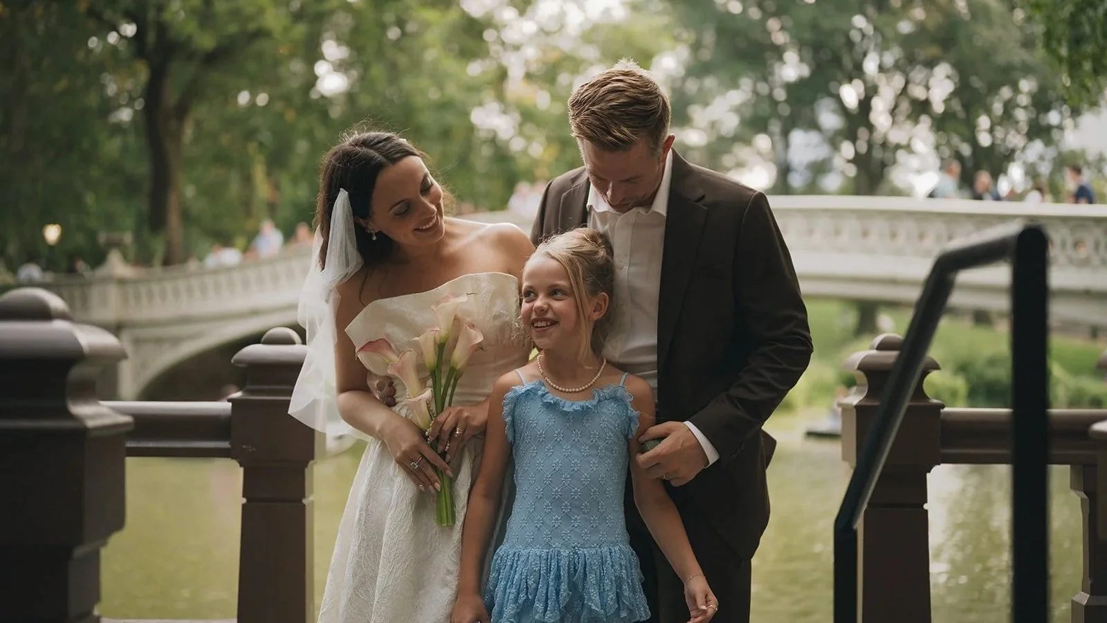 A bride in a white wedding dress holding a bouquet of flowers, smiling at a young girl in a blue dress, with a man in a suit looking at the girl. They are outdoors near a bridge and water, with trees in the background.
