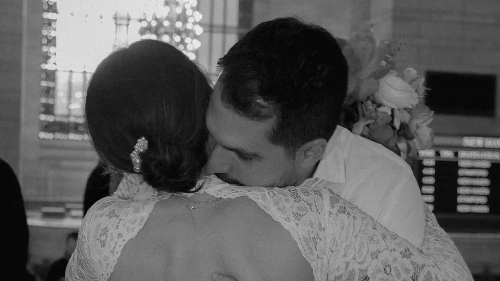 A black and white photo of a man and woman embracing, possibly kissing, at a festive event with trees decorated with lights in the background.