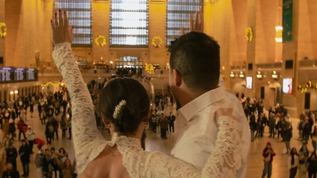 A couple dressed in formal attire, a woman with an updo hairstyle and a man with short dark hair, holding hands and looking at each other in a busy train station with a high ceiling and many people.