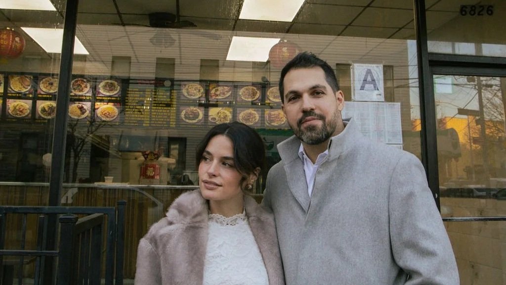 A man and woman standing outside a restaurant with a menu written on the window in the background.