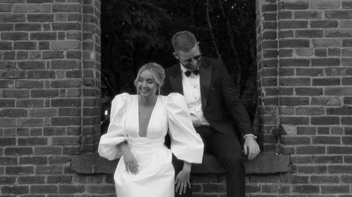 A smiling woman in a white wedding dress with puffed sleeves seated on a brick ledge, next to a man in a black tuxedo with a bowtie, sitting on the same ledge, both by a brick wall with greenery in the background.