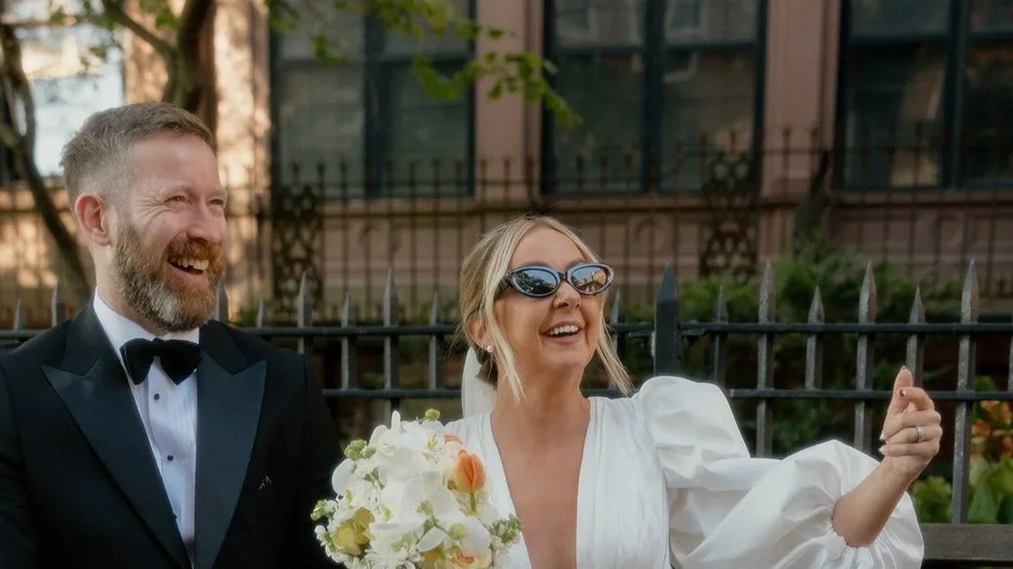 Woman in white dress and sunglasses taking a selfie with man in a tuxedo, holding a bouquet of flowers, both smiling.