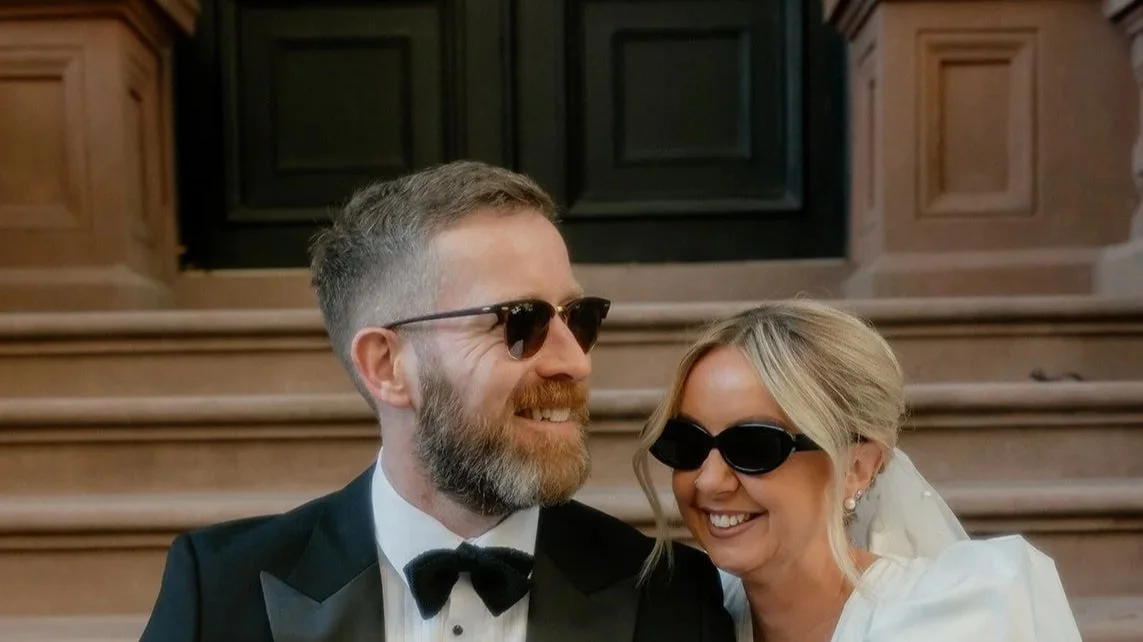 A man and woman dressed in wedding attire wearing sunglasses, smiling and standing close together on a staircase with wooden paneling.