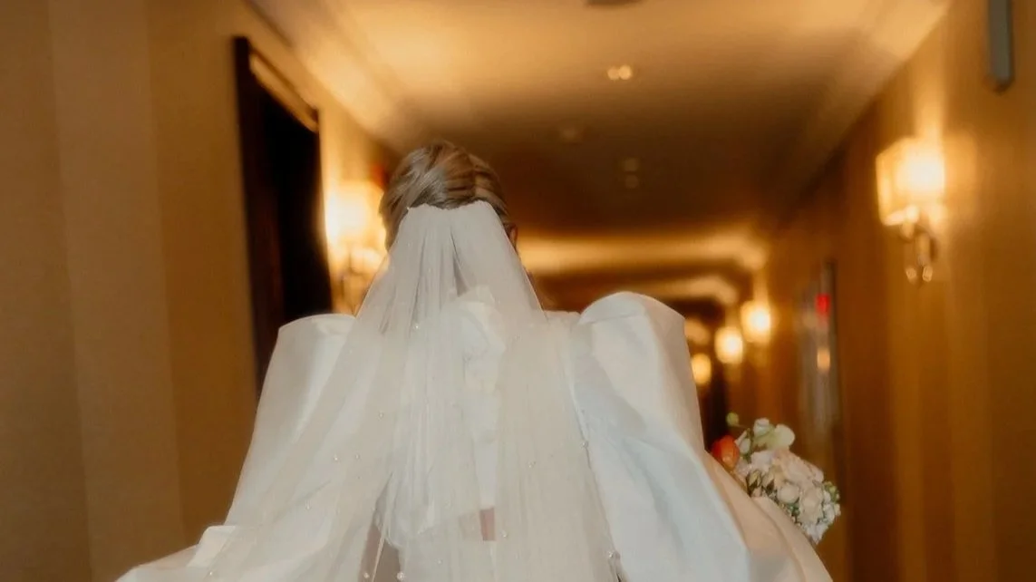 Bride in a wedding dress and veil holding a bouquet walking down a hotel corridor.