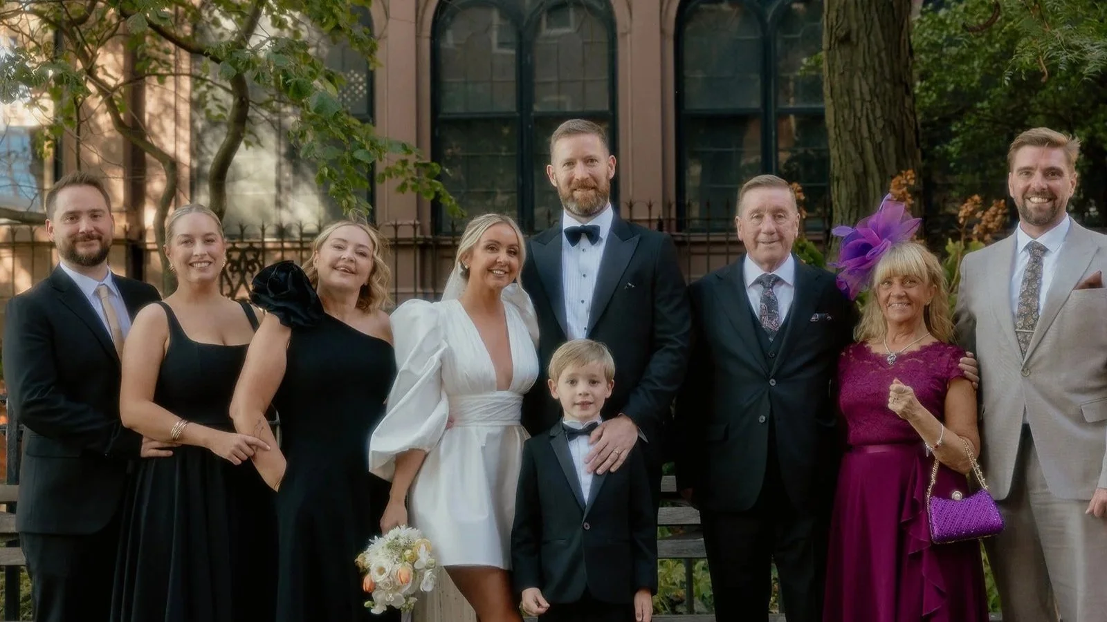 Group of people dressed in wedding attire, standing outdoors in front of a building and trees, celebrating a wedding.