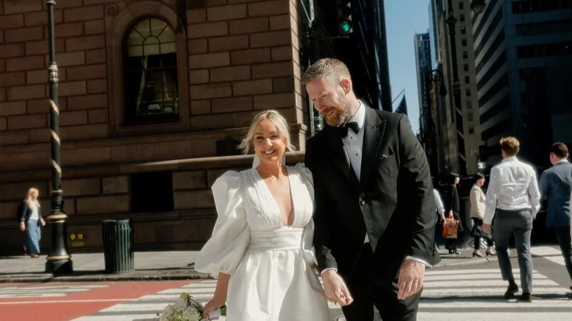 A bride and groom walking on a city street, smiling, with buildings and pedestrians in the background.