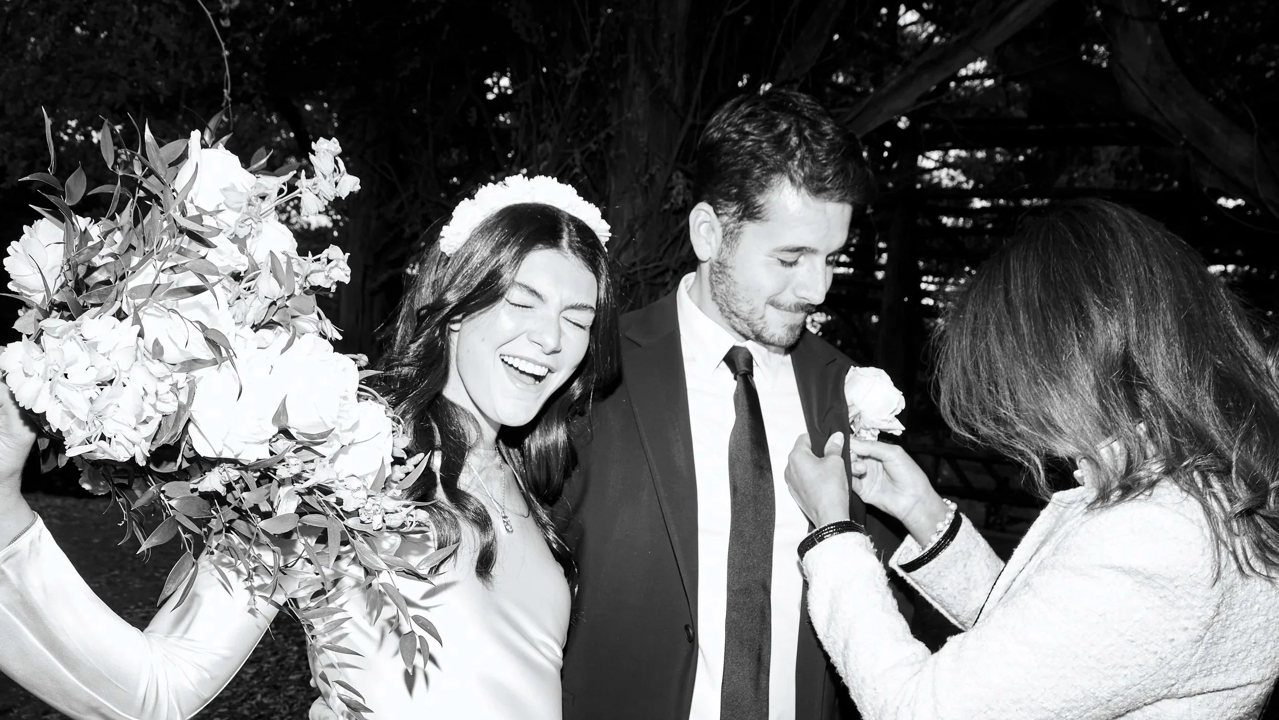 A black-and-white photo showing a woman with long dark hair smiling with eyes closed, wearing a flower crown. She is in a wedding dress and holding a large bouquet of flowers. Next to her is a man with short dark hair and beard, wearing a suit and ti