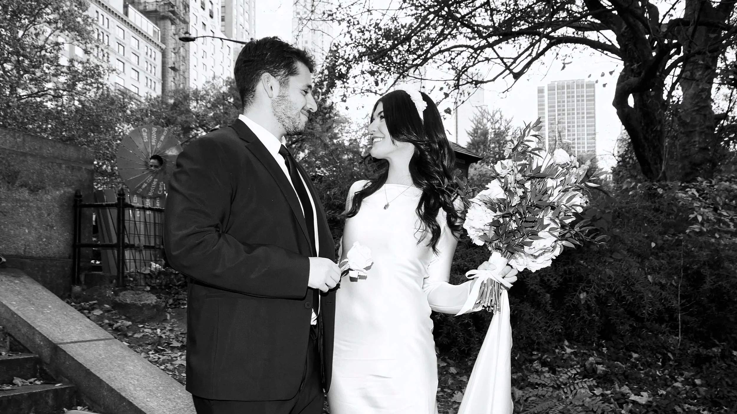 A black and white photo of a couple in a park, with the man in a suit and the woman in a wedding dress holding a bouquet of flowers, smiling at each other surrounded by trees and urban buildings in the background.