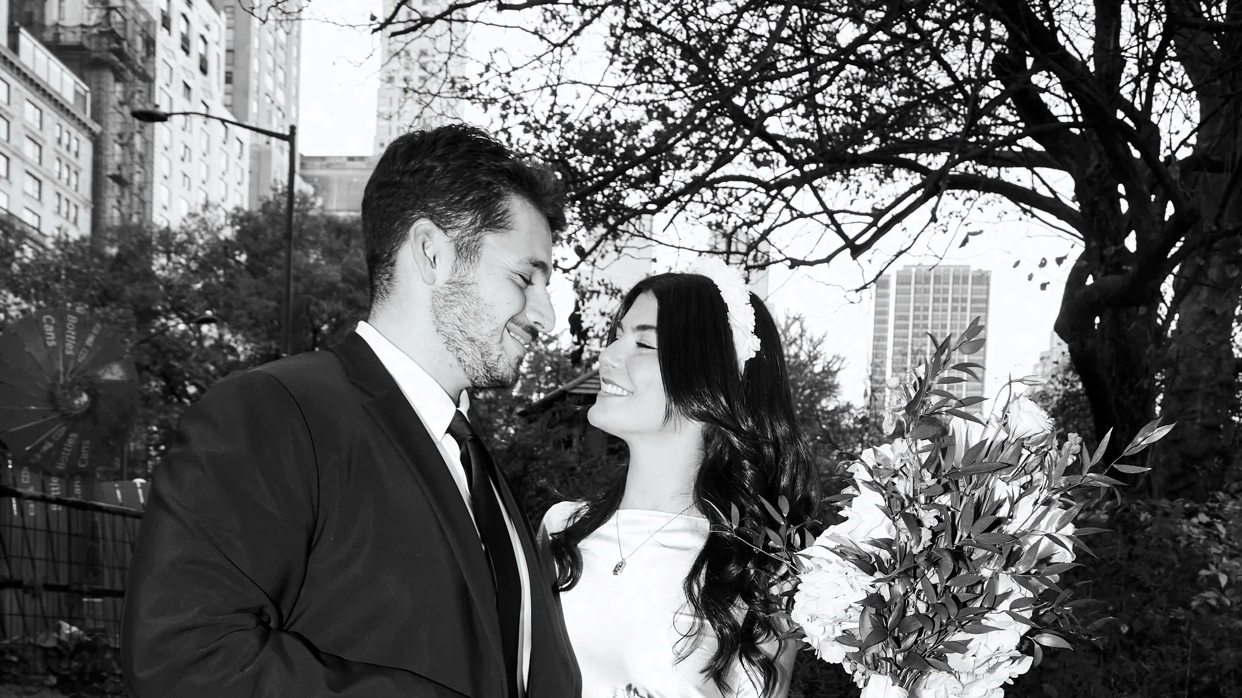 Black and white photo of a smiling couple facing each other with eyes closed, standing outdoors among trees and city buildings in the background. The man is wearing a suit and the woman has long dark hair and is holding a bouquet of flowers.