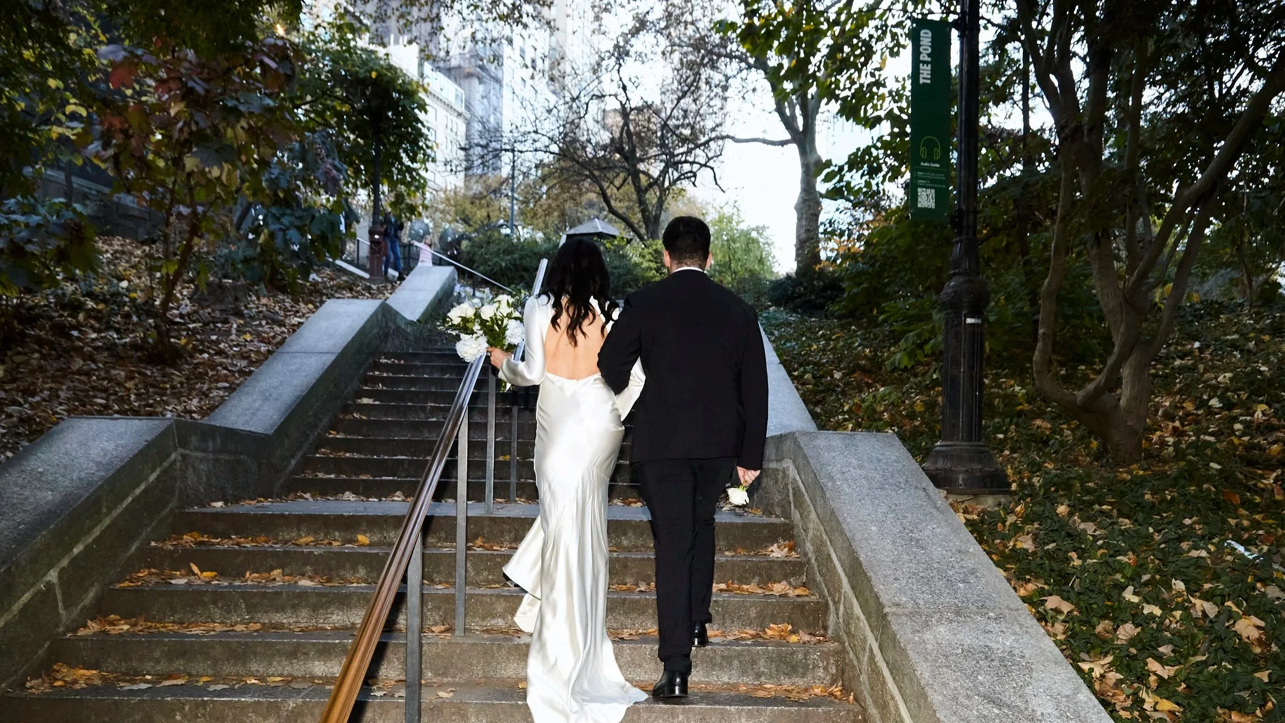 A bride and groom walking up outdoor stairs in park, holding hands, bride with white dress holding bouquet, groom in dark suit, autumn leaves, background trees, street lamp.
