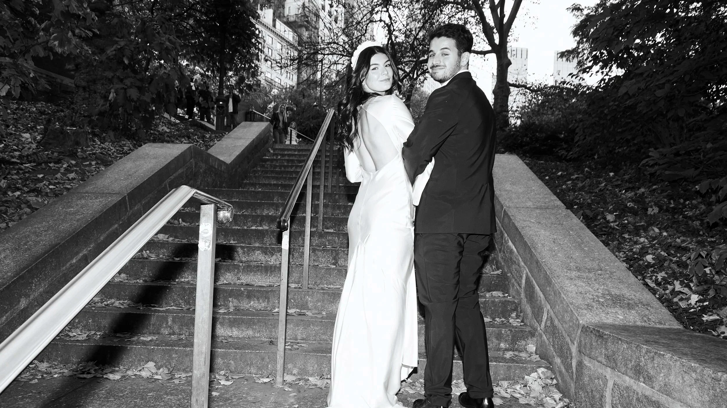 A couple in wedding attire standing on outdoor stairs, surrounded by trees and city buildings, with other people in the background.